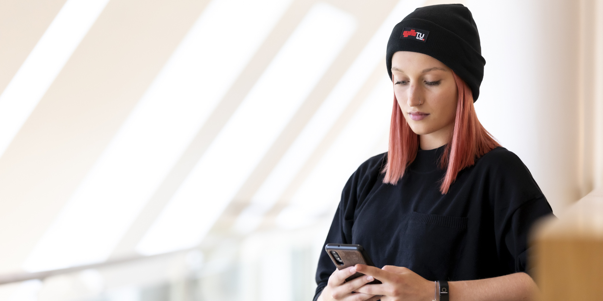 Eine Frau mit einer schwarzen Haube und einem schwarzen T-Shirt blickt konzentriert auf ein Smartphone.